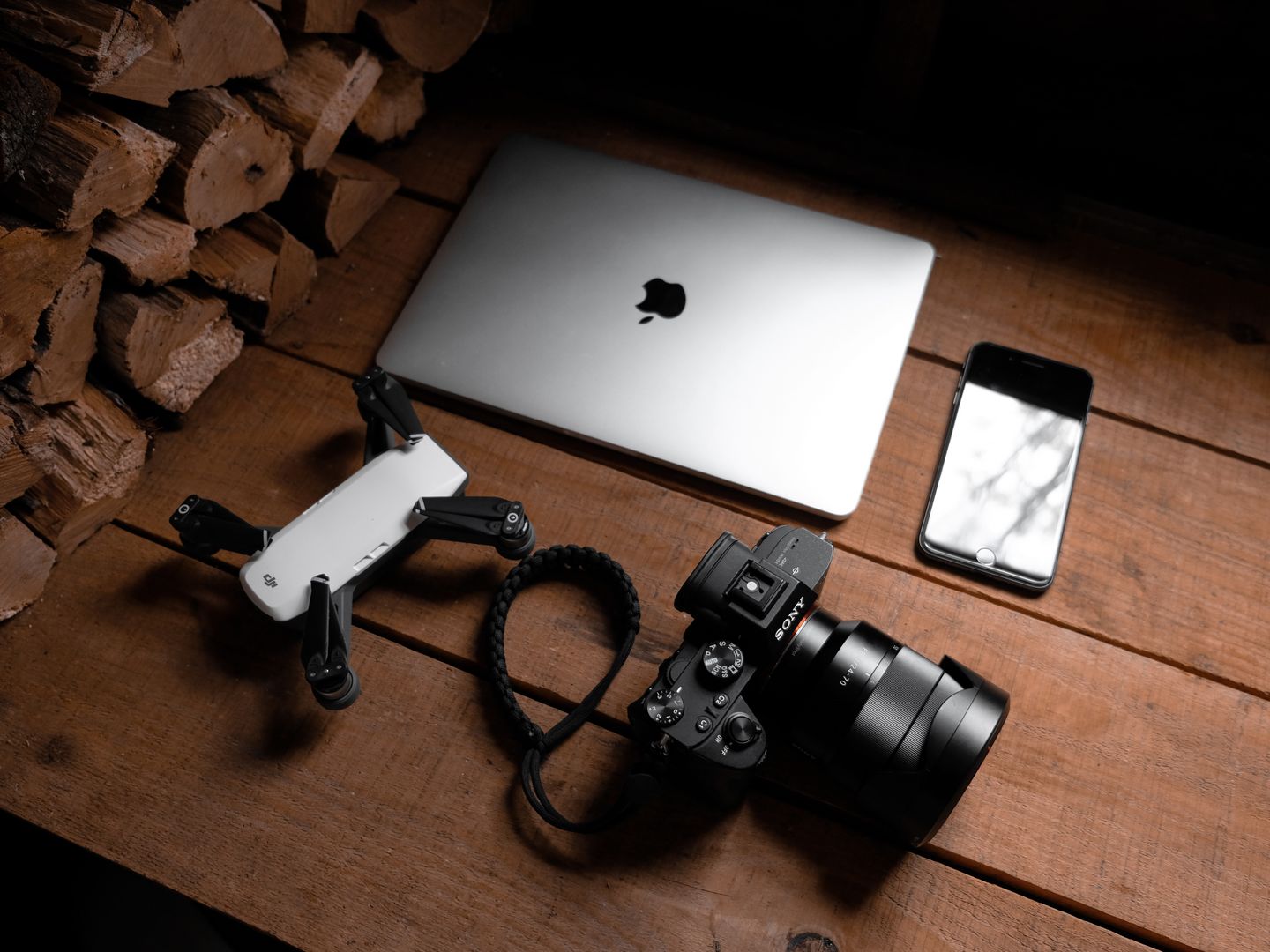 A flat lay of modern technology devices including a laptop, camera, drone, and smartphone on a wooden table.